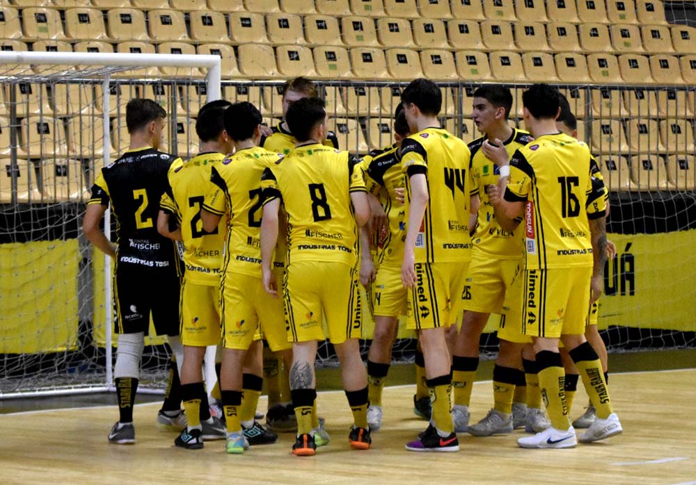 Atletas da base do Jaraguá Futsal, uniformizados de amarelo e preto, reunidos em quadra durante um jogo, com o goleiro adversário de preto ao lado