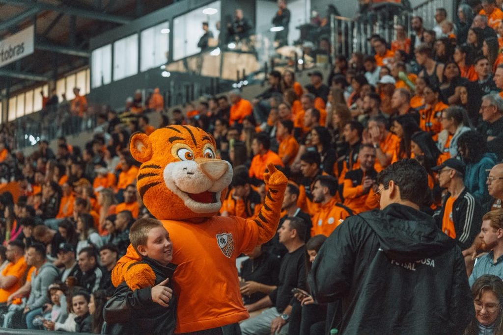 Mascote da ACBF abraça uma criança em meio à torcida de camisas laranja, no Centro Municipal de Eventos, em Carlos Barbosa